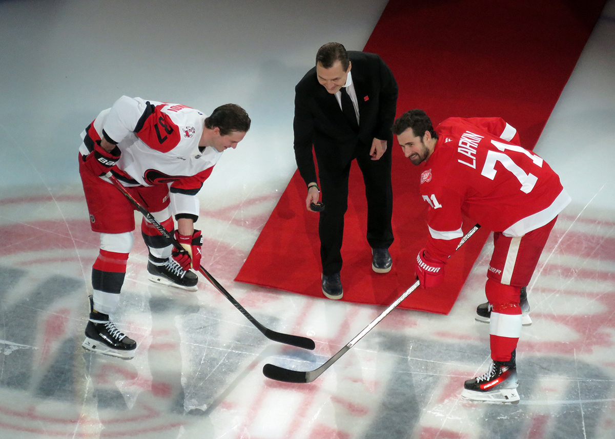 Andrei Svechnikov of the Carolina Hurricanes and Dylan Larkin of the Detroit Red Wings prepare for a ceremonial faceoff with Sergei Fedorov as part of Fedorov's jersey retirement ceremony.